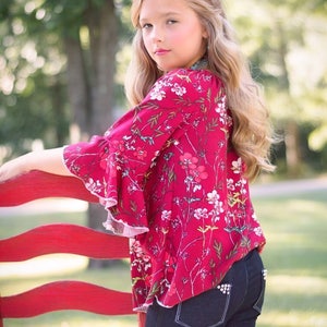 May include: A young girl wearing a red floral blouse with ruffled sleeves and dark blue jeans. She is standing next to a red wooden chair.