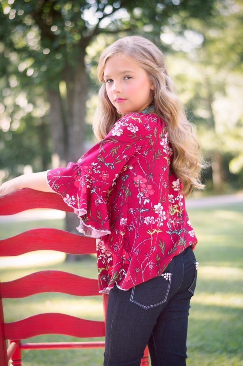 May include: A young girl wearing a red floral blouse with ruffled sleeves and dark blue jeans. She is standing next to a red wooden chair.