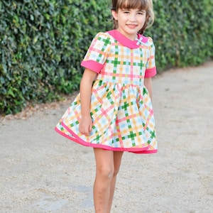 May include: A young girl wearing a pink and white gingham dress with a pink collar and a bow. She is wearing gold sandals and standing on a gravel path.