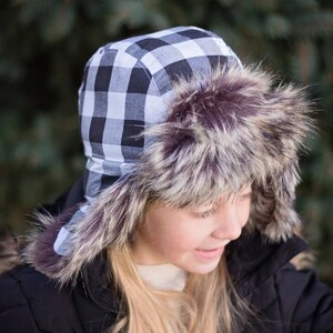 May include: A black and white checkered trapper hat with a faux fur brim. The hat is worn by a person with long blonde hair.