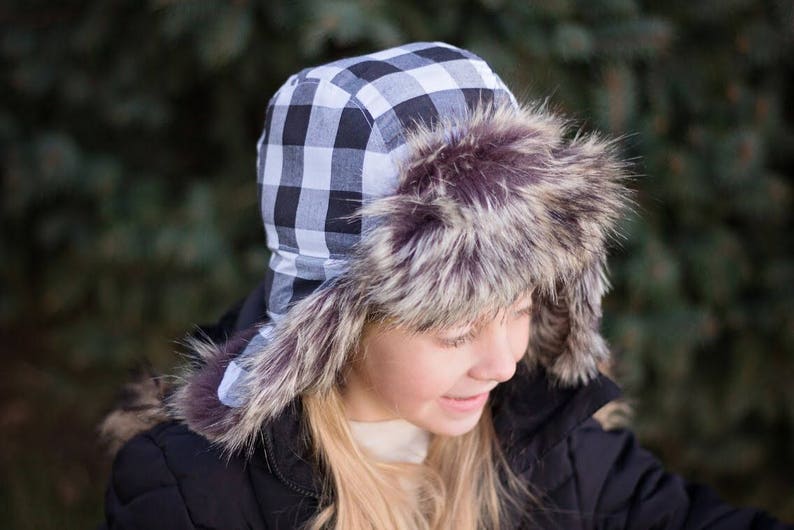 May include: A black and white checkered trapper hat with a faux fur brim. The hat is worn by a person with long blonde hair.