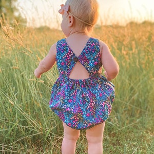 May include: A toddler wearing a blue and pink floral print romper with a cutout back. The romper has a button closure at the back. The toddler is standing in a field of tall grass.