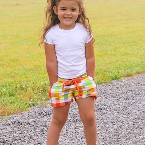 May include: A young girl wearing a white t-shirt and colorful plaid shorts stands on a gravel path. She has long brown hair with a yellow scrunchie and is smiling at the camera.