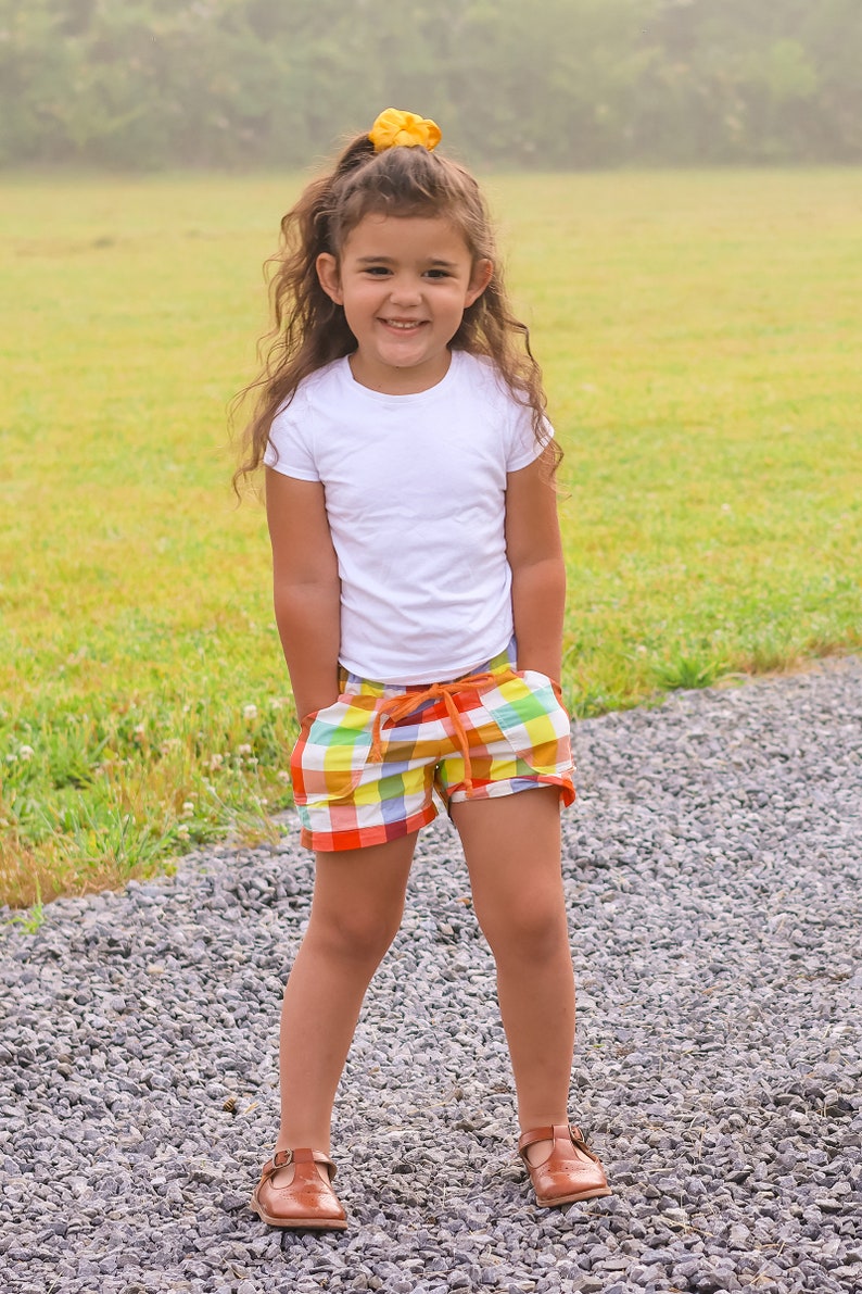 May include: A young girl wearing a white t-shirt and colorful plaid shorts stands on a gravel path. She has long brown hair with a yellow scrunchie and is smiling at the camera.
