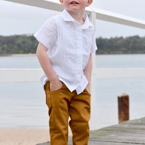 Op de afbeelding: Een jonge jongen in een wit linnen shirt en een bruine broek staat op een houten pier. Hij kijkt omhoog en naar rechts.