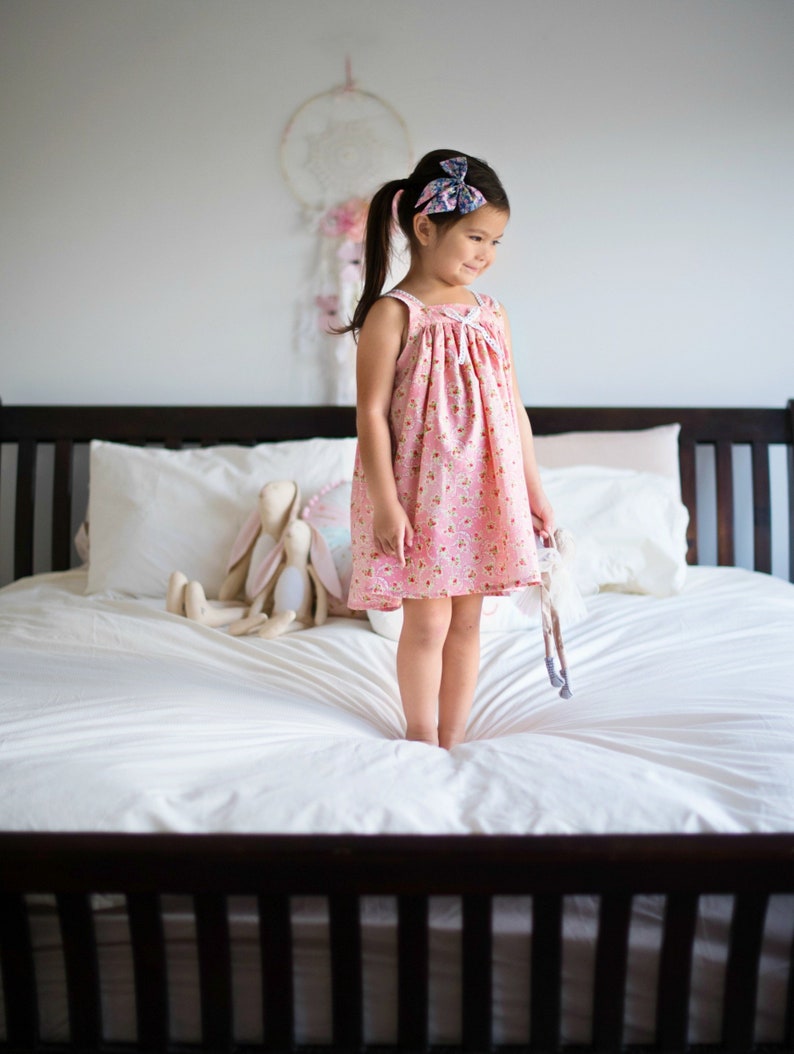 Pu&ograve; includere: Una bambina con un vestito rosa a fiori sta in piedi su un letto bianco con un peluche. Indossa un fiocco blu e viola nei capelli.