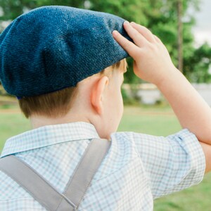 May include: A young boy wearing a blue and white plaid shirt with suspenders and a blue newsboy cap. He is looking off to the side and has his hand on his cap.