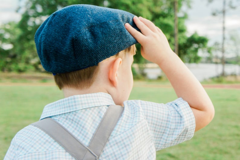 May include: A young boy wearing a blue and white plaid shirt with suspenders and a blue newsboy cap. He is looking off to the side and has his hand on his cap.