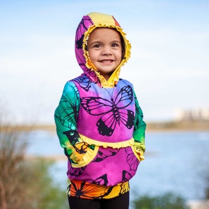Puede incluir: Un niño pequeño con una sudadera con capucha de colores con estampado de mariposas. La sudadera tiene un ribete de volantes amarillo y bolsillos. El niño sonríe y mira a la cámara.