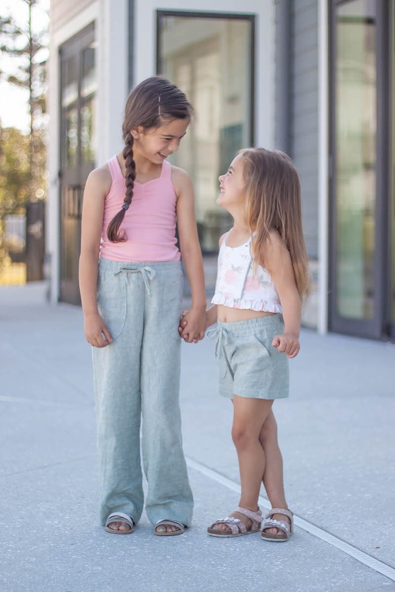 May include: Two girls wearing casual summer outfits stand outside. The girl on the left is wearing a pink tank top and light blue wide-leg pants. The girl on the right is wearing a white floral crop top and light blue shorts.