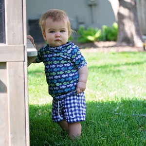 Puede incluir: Un niño pequeño con una camisa de manga corta azul y verde con un patrón de pescado y pantalones cortos de cuadros azules y blancos está de pie en un área cubierta de hierba.