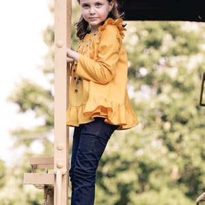 May include: A young girl wearing a yellow ruffled blouse and blue jeans stands on a metal platform. She is looking at the camera.