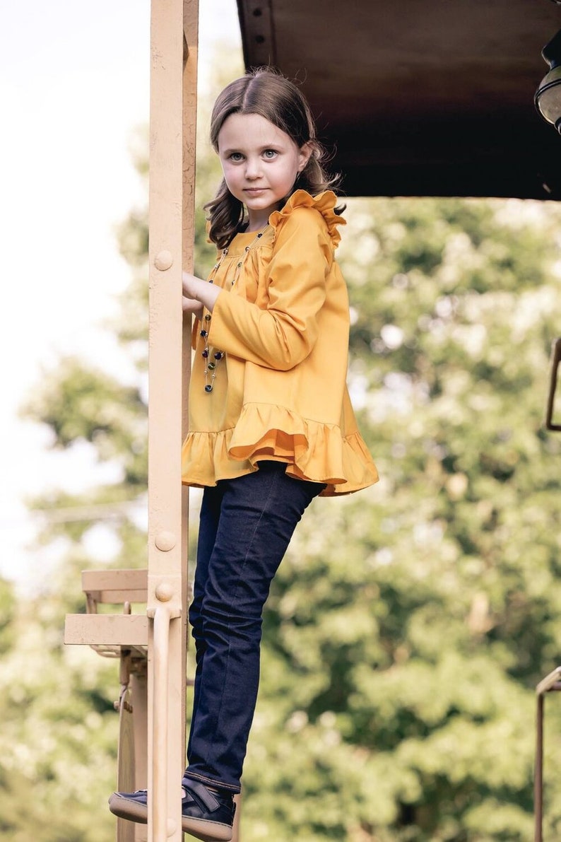 May include: A young girl wearing a yellow ruffled blouse and blue jeans stands on a metal platform. She is looking at the camera.