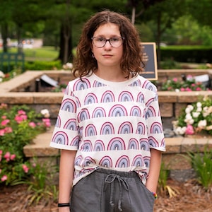 May include: A young person wearing a white t-shirt with a rainbow pattern and gray shorts. They are wearing glasses and have curly brown hair.