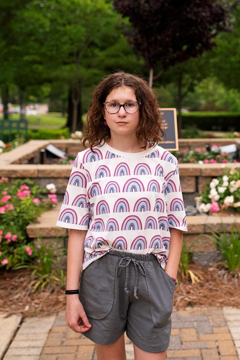 May include: A young person wearing a white t-shirt with a rainbow pattern and gray shorts. They are wearing glasses and have curly brown hair.