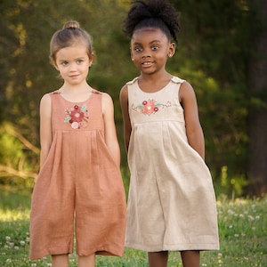 May include: Two young girls wearing linen dresses with embroidered floral details. The girl on the left is wearing a rust-colored dress with a floral design on the chest. The girl on the right is wearing a beige dress with a floral design on the chest. Both girls are standing in a grassy field.