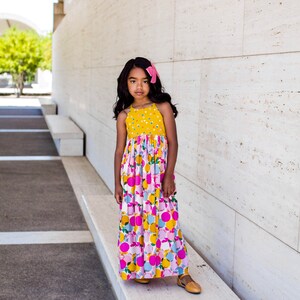 May include: A young person wearing a yellow and pink floral maxi dress with a pink bow in their hair. They are standing in front of a white wall.