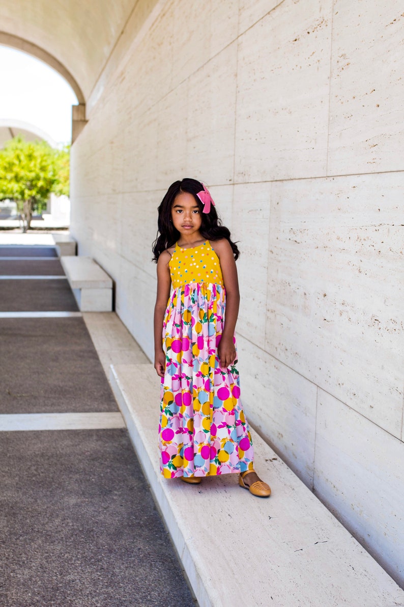 May include: A young person wearing a yellow and pink floral maxi dress with a pink bow in their hair. They are standing in front of a white wall.