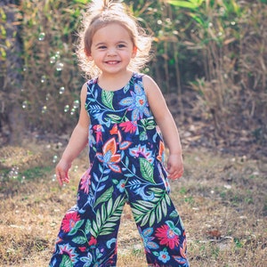 May include: A young girl wearing a blue and floral print jumpsuit with a wide leg. She is standing in a grassy field with green foliage in the background.
