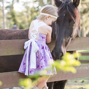 May include: A young girl in a purple and white floral dress with a white bow is standing next to a black horse. She is leaning against a wooden fence and petting the horse's head.