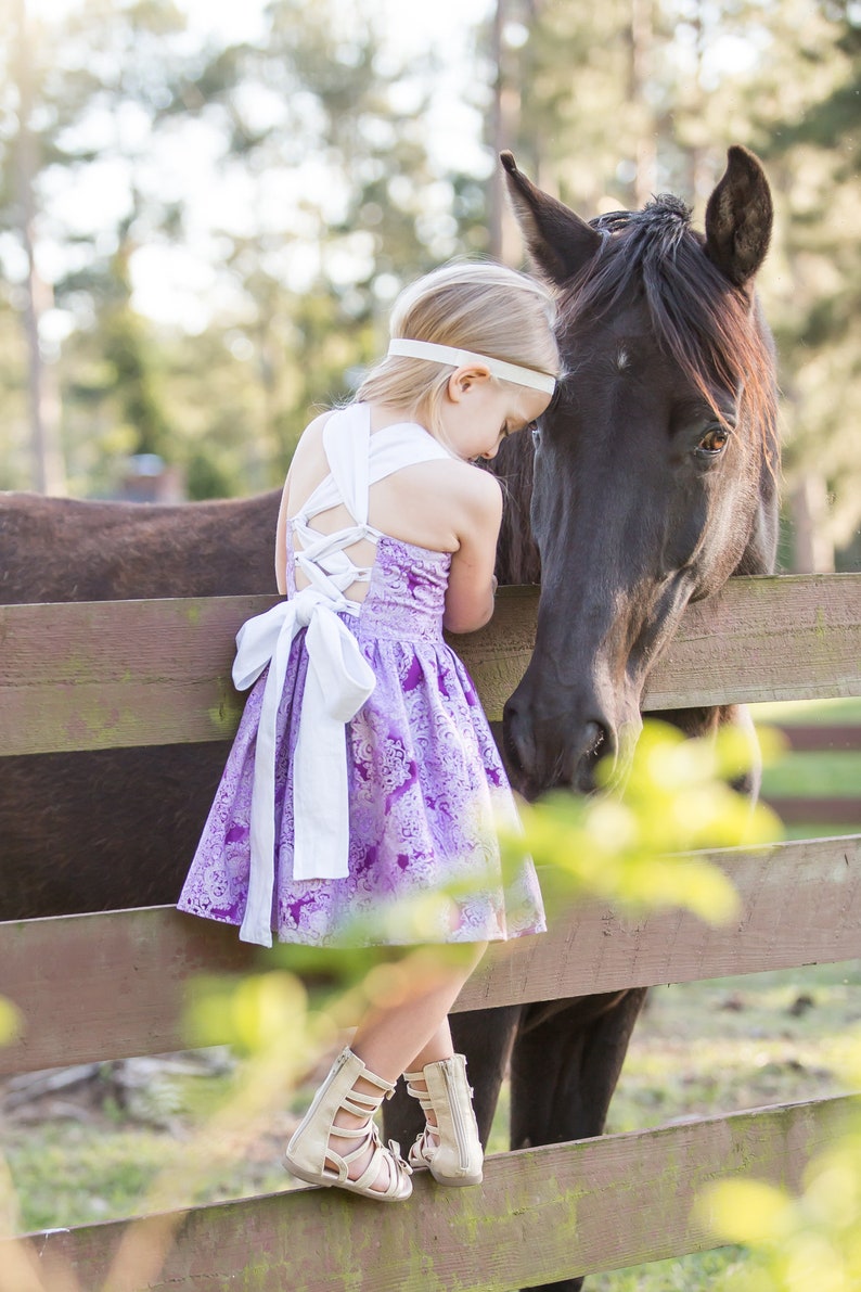 May include: A young girl in a purple and white floral dress with a white bow is standing next to a black horse. She is leaning against a wooden fence and petting the horse's head.