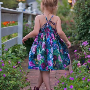May include: A young girl wearing a floral print dress with a purple flower hair clip walks on a brick path lined with purple flowers.