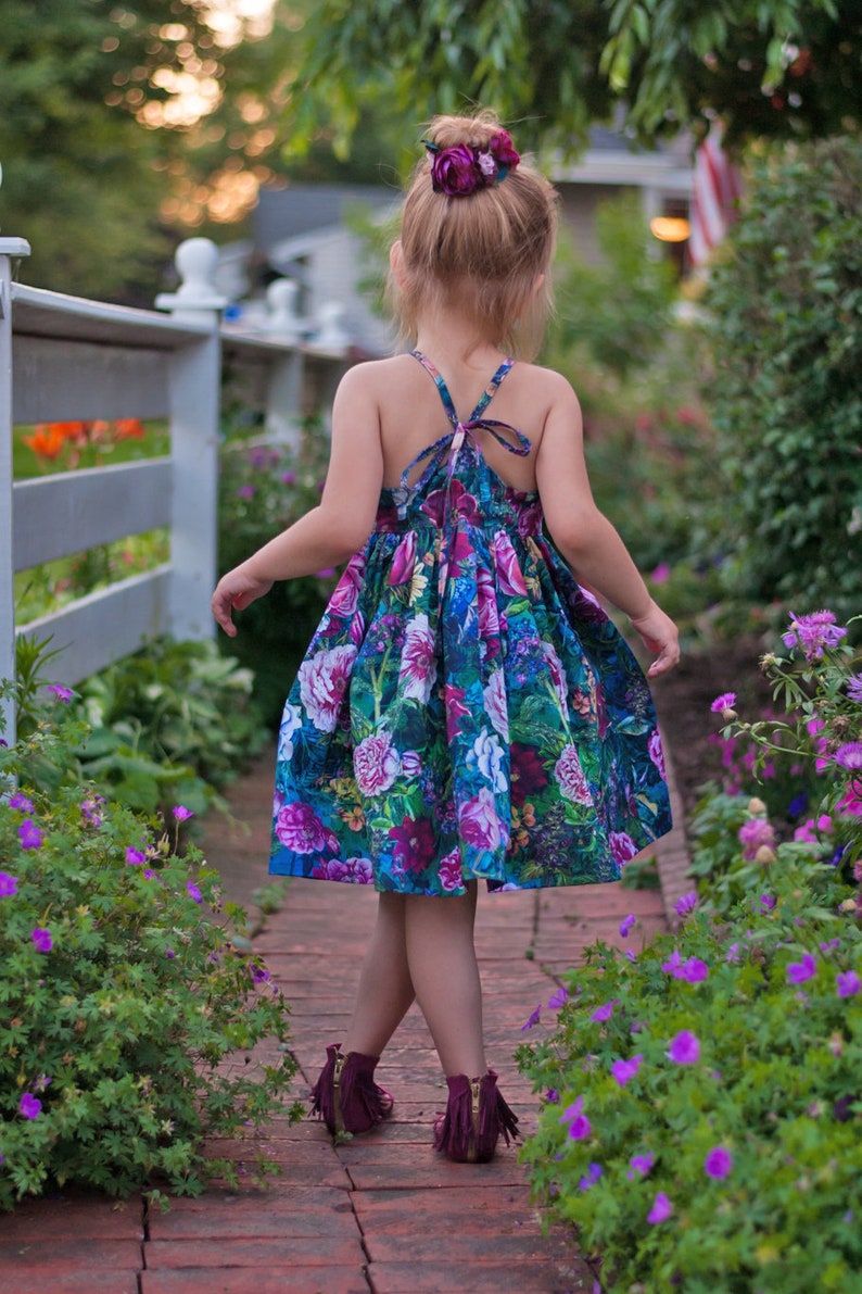 May include: A young girl wearing a floral print dress with a purple flower hair clip walks on a brick path lined with purple flowers.