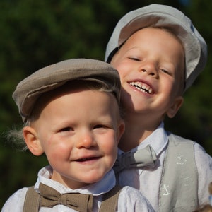 May include: Two young boys wearing white shirts and brown bow ties. The boy on the left is wearing a brown cap and suspenders. The boy on the right is wearing a light gray cap and a vest with anchor designs. Both boys are smiling.