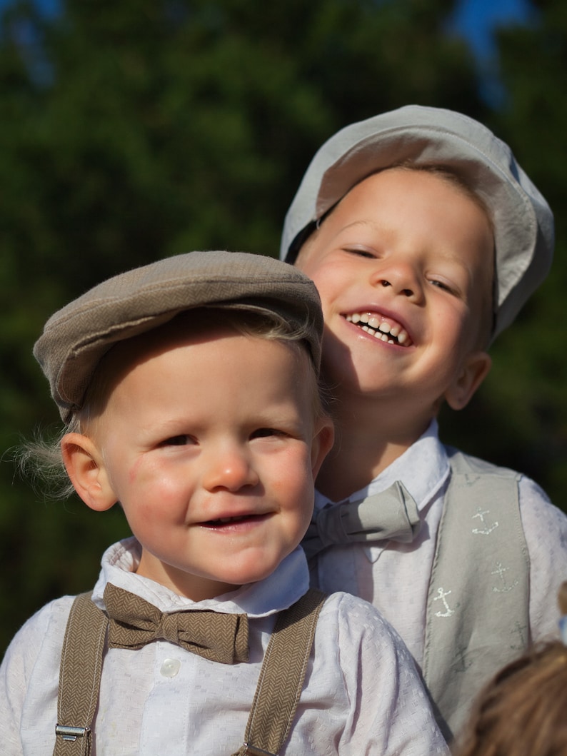 May include: Two young boys wearing white shirts and brown bow ties. The boy on the left is wearing a brown cap and suspenders. The boy on the right is wearing a light gray cap and a vest with anchor designs. Both boys are smiling.