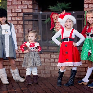 May include: Four girls wearing festive Christmas dresses. The girls are standing in front of a brick fireplace with a wreath and presents. The dresses are red, green, black and white with various Christmas-themed designs.