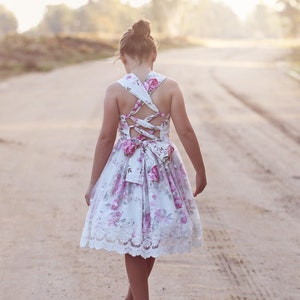 May include: A young person wearing a white dress with pink floral print and lace trim. The dress has a criss-cross back design with a bow at the waist.