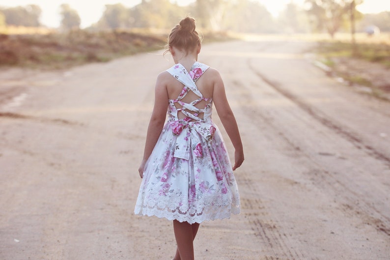 May include: A young person wearing a white dress with pink floral print and lace trim. The dress has a criss-cross back design with a bow at the waist.