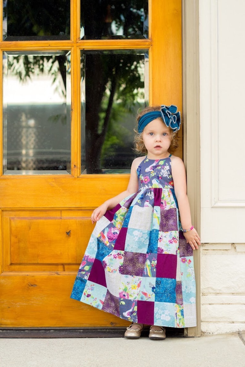 May include: A young girl wearing a teal headband with a bow and a colorful patchwork dress stands in front of a wooden door.