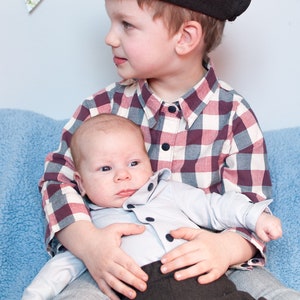 May include: A young boy wearing a black and white plaid shirt and a black newsboy cap holds a baby wearing a light blue button-down shirt and dark gray pants.