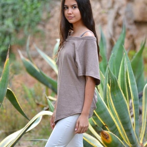 May include: A young person wearing a brown off-the-shoulder top and white pants stands in front of a large green plant with yellow stripes.