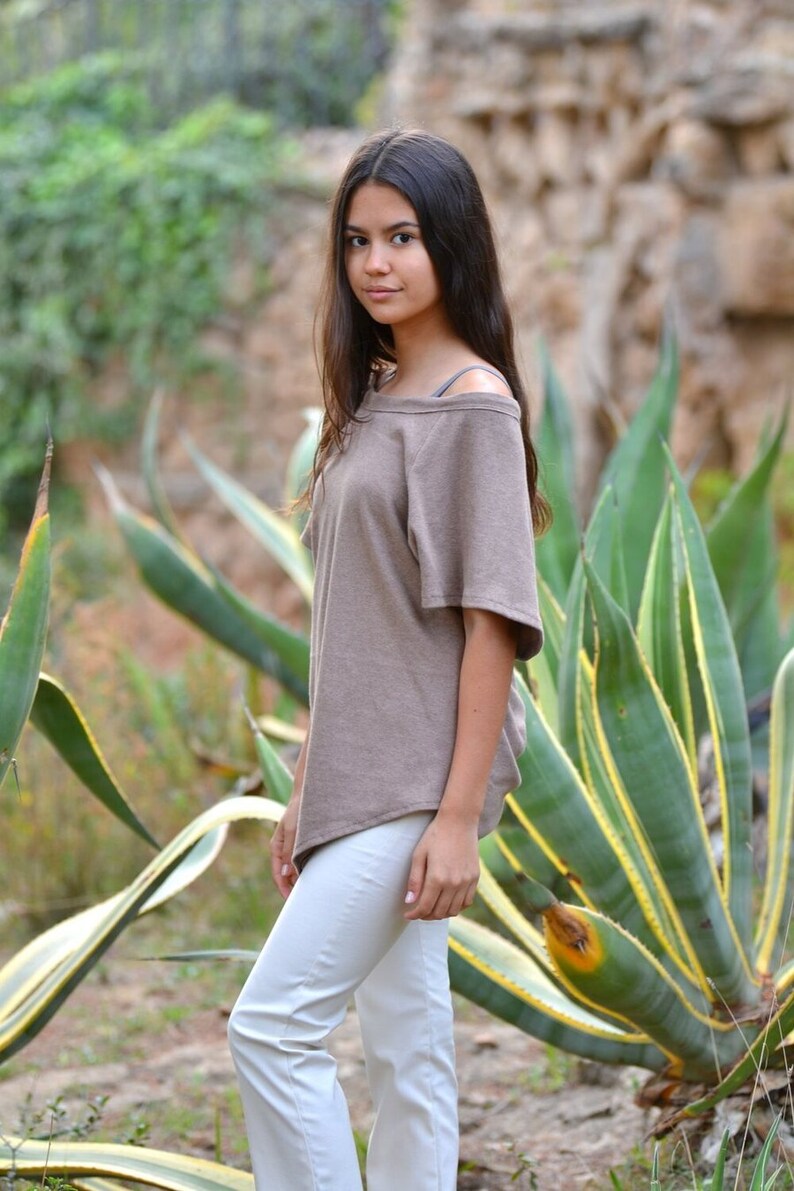 May include: A young person wearing a brown off-the-shoulder top and white pants stands in front of a large green plant with yellow stripes.