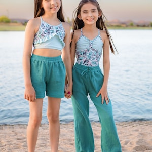 May include: Two girls wearing teal swimsuits and matching teal pants stand on a sandy beach. The girl on the left is wearing a light blue ruffle top with a floral print. The girl on the right is wearing a teal swimsuit with a floral print. Both girls are smiling.