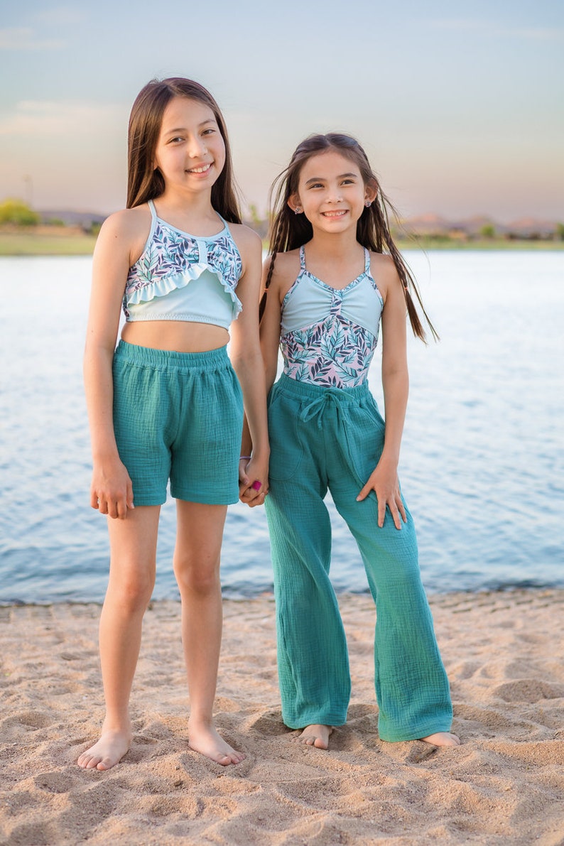 May include: Two girls wearing teal swimsuits and matching teal pants stand on a sandy beach. The girl on the left is wearing a light blue ruffle top with a floral print. The girl on the right is wearing a teal swimsuit with a floral print. Both girls are smiling.