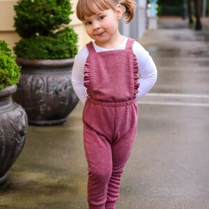 May include: A young girl wearing a pink ruffled jumpsuit and black shoes. She is standing on a sidewalk in front of a building.