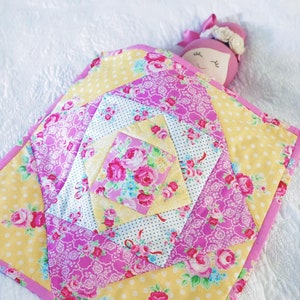 May include: A pink and yellow patchwork quilt with a floral pattern. The quilt is folded in half and has a doll lying on top of it.