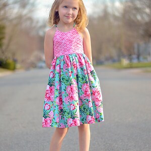 May include: A young girl wearing a pink and blue floral dress with white sandals. The dress has a pink bodice with a white pattern and a blue skirt with a floral pattern.