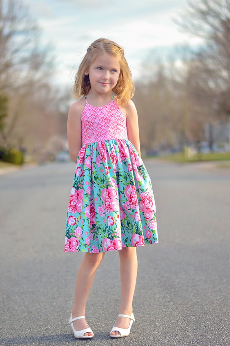 May include: A young girl wearing a pink and blue floral dress with white sandals. The dress has a pink bodice with a white pattern and a blue skirt with a floral pattern.