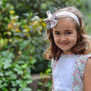 May include: A young girl with long brown hair smiles at the camera. She is wearing a white dress with a floral pattern and a floral headband. The background is a blurred image of green foliage.