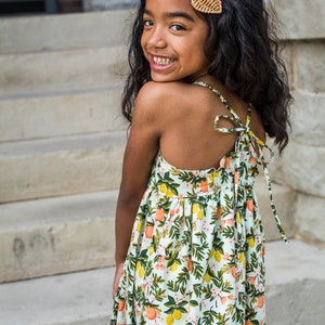 May include: A young girl wearing a white dress with a lemon and orange print. The dress has a tie at the back and the girl is smiling. She has a brown heart-shaped hair clip in her hair.