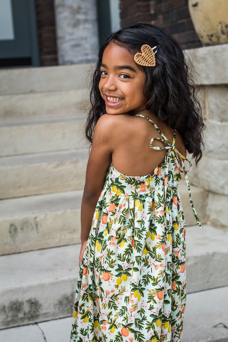 May include: A young girl wearing a white dress with a lemon and orange print. The dress has a tie at the back and the girl is smiling. She has a brown heart-shaped hair clip in her hair.