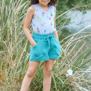 May include: A young person wearing a light gray tank top with a pineapple print, teal blue shorts, a straw hat, and blue sandals. The person is standing in a grassy area with tall green reeds.