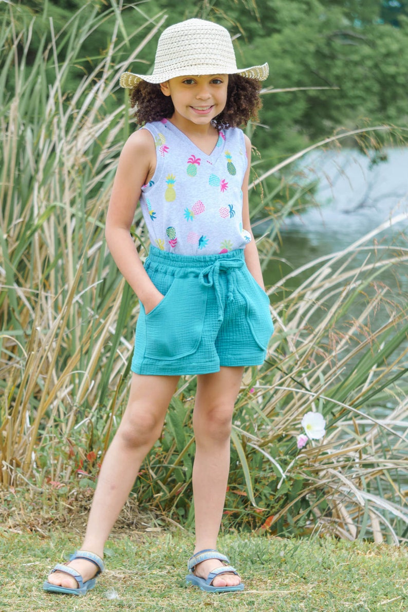 May include: A young person wearing a light gray tank top with a pineapple print, teal blue shorts, a straw hat, and blue sandals. The person is standing in a grassy area with tall green reeds.