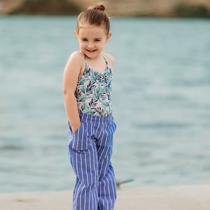 May include: A young girl wearing a floral print tank top and blue and white striped pants stands on a sandy surface with a body of water in the background.