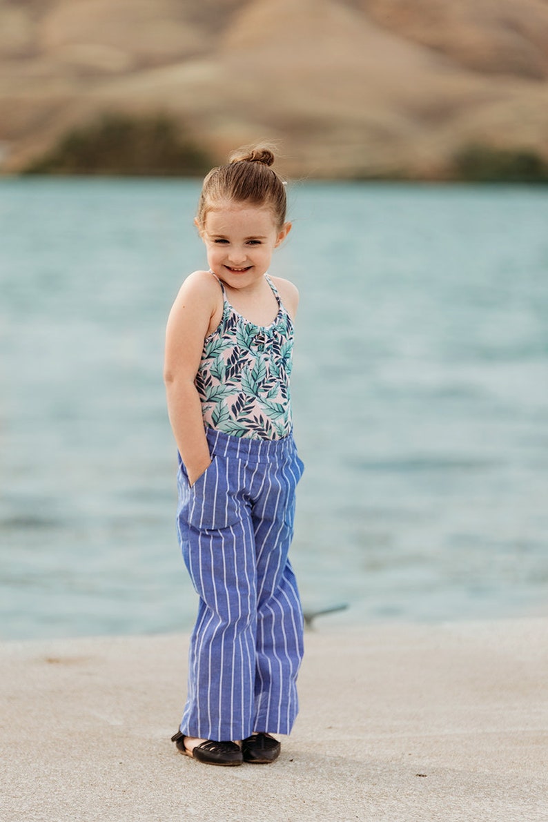 May include: A young girl wearing a floral print tank top and blue and white striped pants stands on a sandy surface with a body of water in the background.