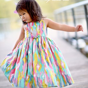 May include: A young girl wearing a colorful, abstract print dress with a beaded necklace. The dress has a gathered skirt and a sleeveless bodice. The girl is standing on a wooden walkway with the text "my china doll" in the background.